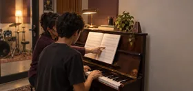 Aluno ao piano de parede com a professora apontando a partitura, bateria ao fundo