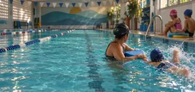 Professora com prancha na borda da raia e criança de touca nadando ao lado, na piscina