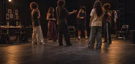 Elenco jovem em roda de ensaio no palco, com refletores acesos e cadeiras encostadas