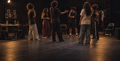 Elenco jovem em roda de ensaio no palco, com refletores acesos e cadeiras encostadas